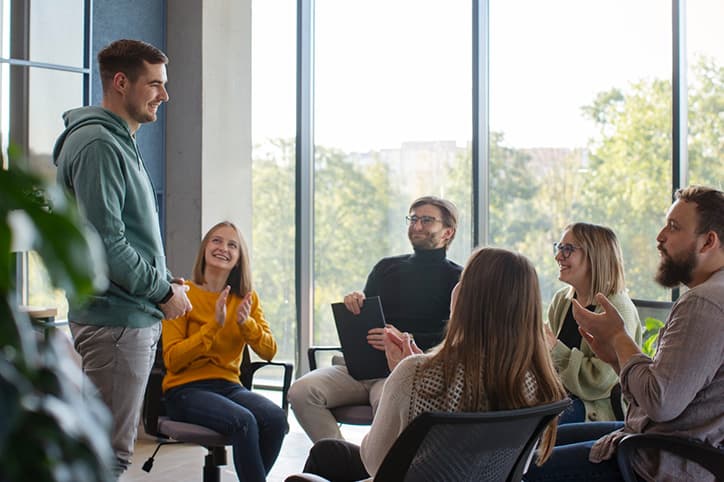 People sitting in a circle in a bright room, listening and applauding as one of them stands to speak.